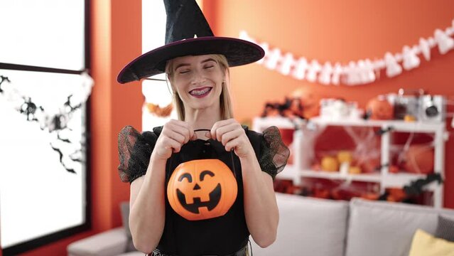 Young blonde woman wearing witch costume holding pumpkin basket at home