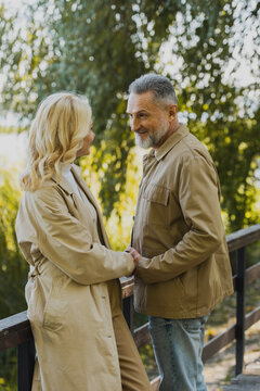 Smiling mature man holding hand of blonde wife while standing on bridge in park.