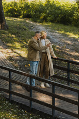 Fototapeta premium Smiling mature man pointing with finger near blonde wife while standing on bridge in park.