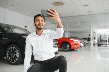 Handsome indian man taking a selfie with car keys to his new automobile at the dealership showroom...