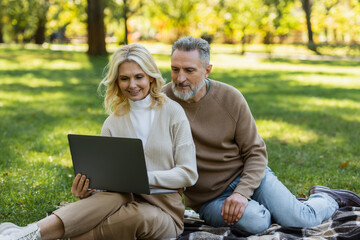 carefree middle aged couple watching movie on laptop and sitting on blanket in park.