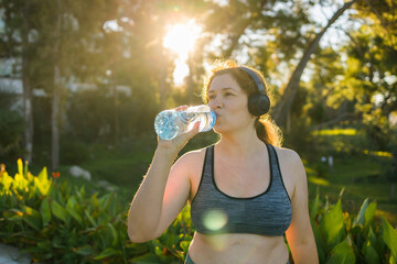 Overweight woman drinking water after jogging in the park. Portrait of young plus-size thirsty woman with a bottle of water outdoors copy space. Sports healthcare and weight loosing fitness and well