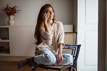 Smiling millennial mixed race female calls by phone, talks, sits on chair looks out window at free space