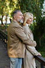 Smiling mature man hugging wife in trench coat while standing on bridge in park.