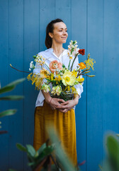 Young woman in flowing yellow skirt and white shirt with beautiful bouquet of peonies in glass vase against the blue background