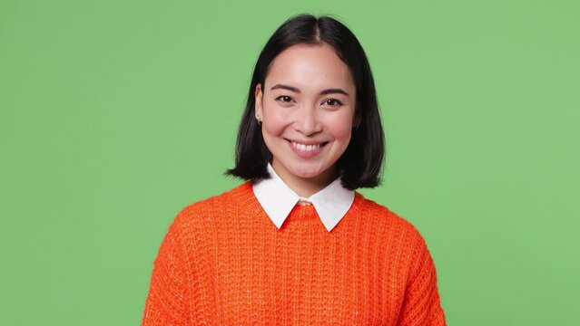 Beautiful smiling charming young woman of Asian ethnicity 20s she wear knitted orange sweater looking camera wink eye blink isolated on plain pastel light green color wall background studio portrait