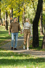 Fototapeta na wymiar full length of stylish middle aged couple holding hands and smiling while walking together in park during springtime.