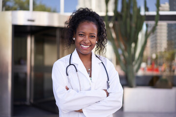 African American female doctor in lab coat with stethoscope outside clinic. Smiling young people medical specialty posing for photo outdoors . Happy and cheerful person with arms crossed.
