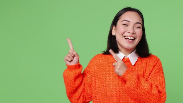 Fun promoter young woman of Asian ethnicity she wear orange sweater pointing indicate index finger aside away on workspace area copy space mock up isolated on plain pastel light green color background