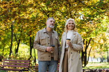 Positive mature couple holding paper cups while walking in park.