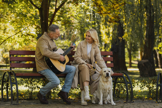 Mature man playing acoustic guitar near smiling wife and labrador in park.