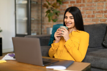 Positive spanish lady watching videos on Internet and drinking coffee at home, looking at laptop screen and smiling