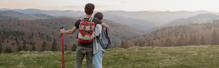Rear of couple in love and hugs standing on mountain peak with walking sticks and watching.