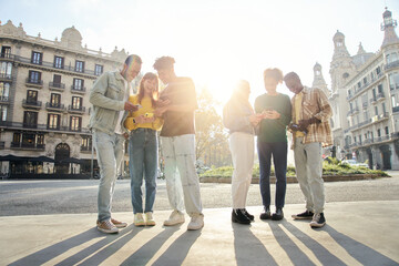 Group of smiling multiracial young people using cell phones. Cheerful students standing with technological devices. Happy university classmates on school trip in a European city. Persons outdoors