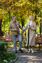 Side view of smiling mature couple holding hands and coffee to go while walking in park.