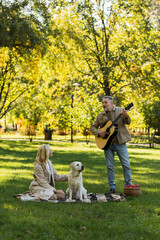Fototapeta premium bearded middle aged man standing near wife and labrador dog while playing acoustic guitar in park.