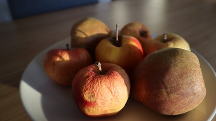 Rotten apples and pears on a plate. Sunlight on the fruits. Plate with the fruits on a wooden table. Plate on a bamboo table. Closeup photo of fruits.