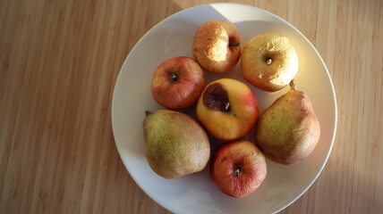 Rotten apples and pears on a plate. Sunlight on the fruits. Plate with the fruits on a wooden table. Plate on a bamboo table. Closeup photo of fruits.
