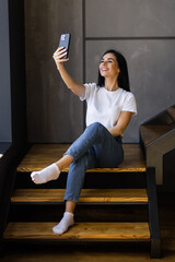 young woman at home in comfortable clothes, making a selfie with smartphone while having a coffee on the stairs of her apartment