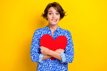 Photo of nervous funky woman dressed blue blouse biting lip embracing red heart isolated yellow...