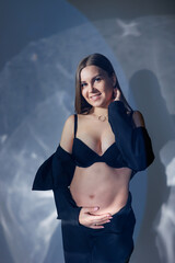 Obraz premium Portrait of a young pregnant woman in a black classic suit. A pregnant woman on a gray background hugs her belly.