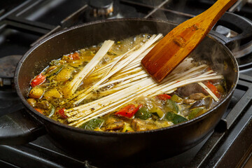 A wooden spatula mixes udon with stewed vegetables in spices in a frying pan.