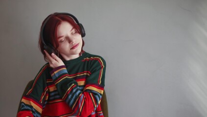 Satisfied girl with red hair 15 years old teenager in headphones enjoys music in a bright striped sweater