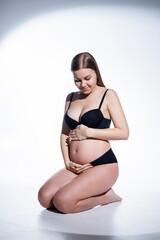 Obraz premium Cute pregnant woman with long hair in black underwear is sitting on a white background. Women's underwear for pregnant women.