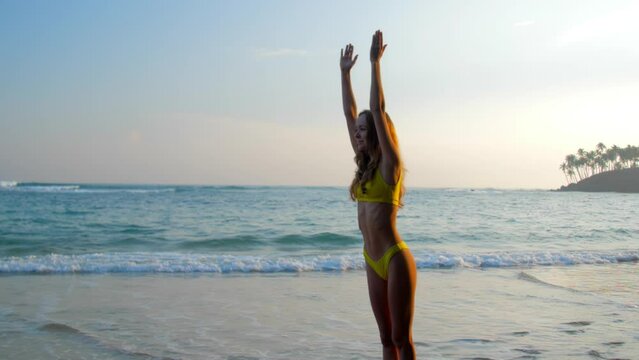 Young woman in bikini does yoga asana raising hands on azure ocean beach at sunrise slow motion. Lady exercises at tropical resort in morning