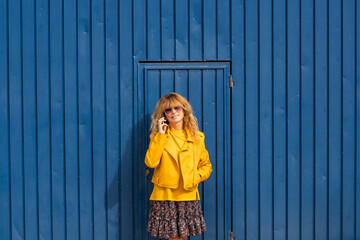 middle age woman with yellow jacket and mobile phone isolated on blue wall in the street