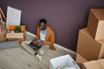 African American man sitting on the floor among boxes and typing on laptop during relocation