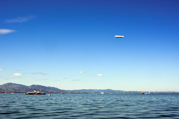 An airship is flying high in the blue sky. Ships and yachts float on the sea under it