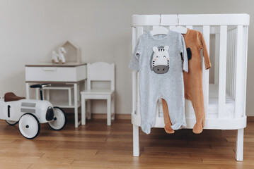 Baby bodysuit with zebra and dog face on hanger on bed, beige background. Cute children's clothes, baby chair, wooden house, retro car in nursery room. Copy space. Scandinavian child room