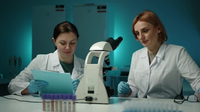 A female scientist looking through a microscope while other one writing something on the tablet. They are in a laboratory. High quality 4k footage 