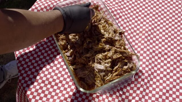 Hands with black gloves shred the meat of a pulled pork with utensils at noon in their garden