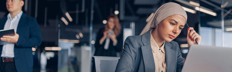 Focused muslim businesswoman in hijab working on computer while sitting in modern office