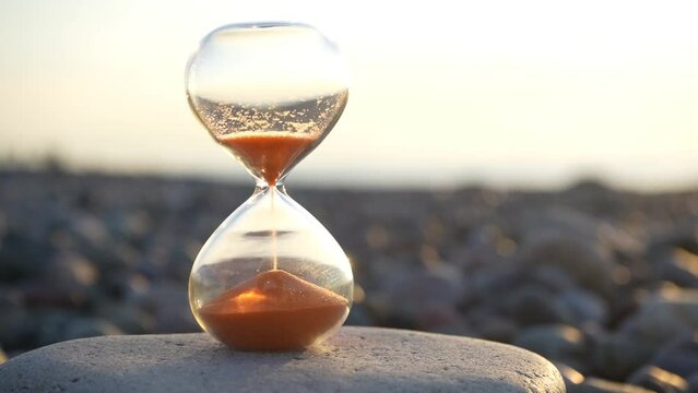 Hourglass on the background of the waves of the sea. Hourglass time on sand and pebble stones coast background. hourglass fall down in water. blurred background, sun set light seen on sunset sky. 