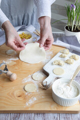 Dough for dumplings, hands, dough, potato filling in white ceramic dishes, wooden spoons, all on a light background.