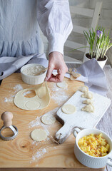 Dough for dumplings, hands, dough, potato filling in white ceramic dishes, wooden spoons, all on a light background.