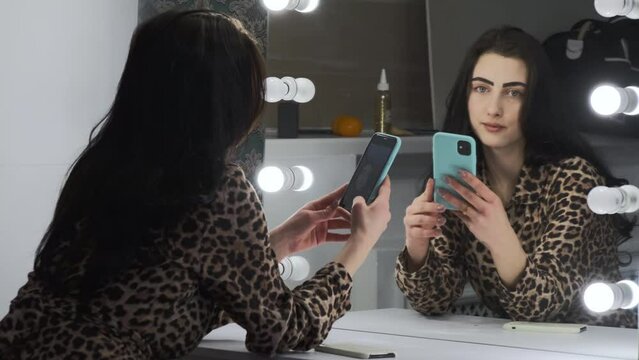 Lady sits and takes photo on phone near illuminated mirror in dressing room. Brunette straightens hair to take good picture on smartphone