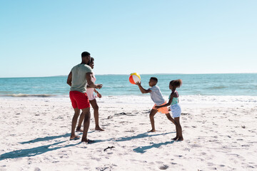 Fototapeta premium African american parents and children playing with ball at beach against beautiful sea and clear sky