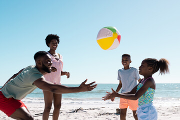 Fototapeta premium Cheerful african american parents and children playing with ball at beach against sea and clear sky