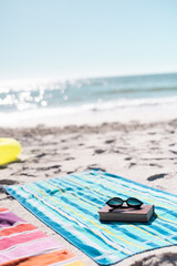 Sunglasses with book placed on blue towel at sandy beach against scenic sea and clear sky