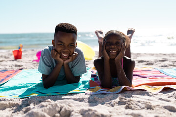 Portrait of african american happy siblings with hands on chins lying on towels at beach against sky