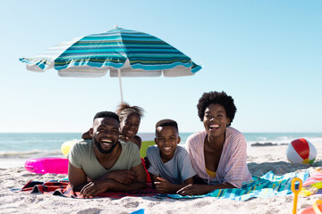 Smiling african american parents and children lying on towels at sandy beach under clear sky