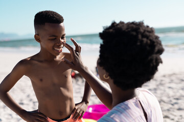 African american mother applying sunscreen on son's nose at beach against clear sky, copy space