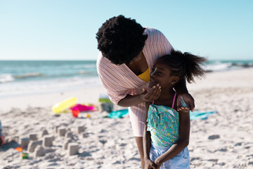 African american mother applying sunscreen on daughter's face at beach against clear sky, copy space