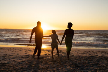 Silhouette african american boy holding mother and father's hands and standing on beach at sunset