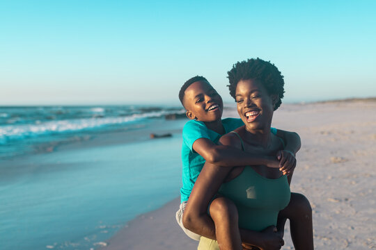 Cheerful african american mother piggybacking son at beach against blue sky during sunset