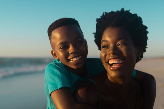 Closeup of happy african american mother piggybacking son at beach against blue sky at sunset